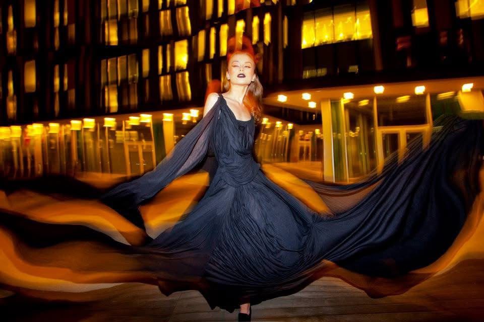 Modella in abito blu notte che vortica in una scena notturna tra luci calde, labbra scure